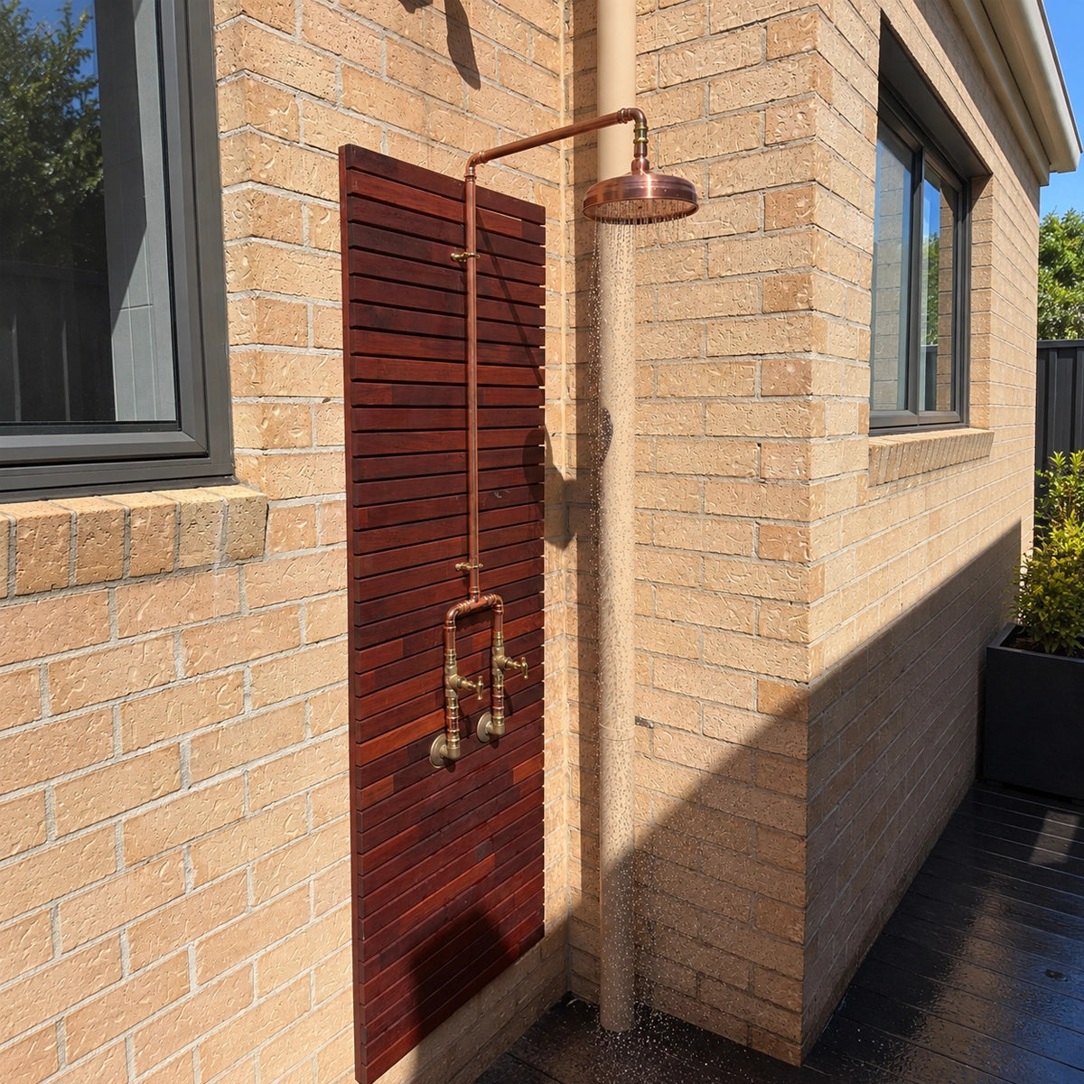 An outdoor wall-mounted shower system installed on a beige brick house exterior, featuring a copper-toned overhead shower arm and head with brass faucets mounted on a dark reddish-brown slatted panel.