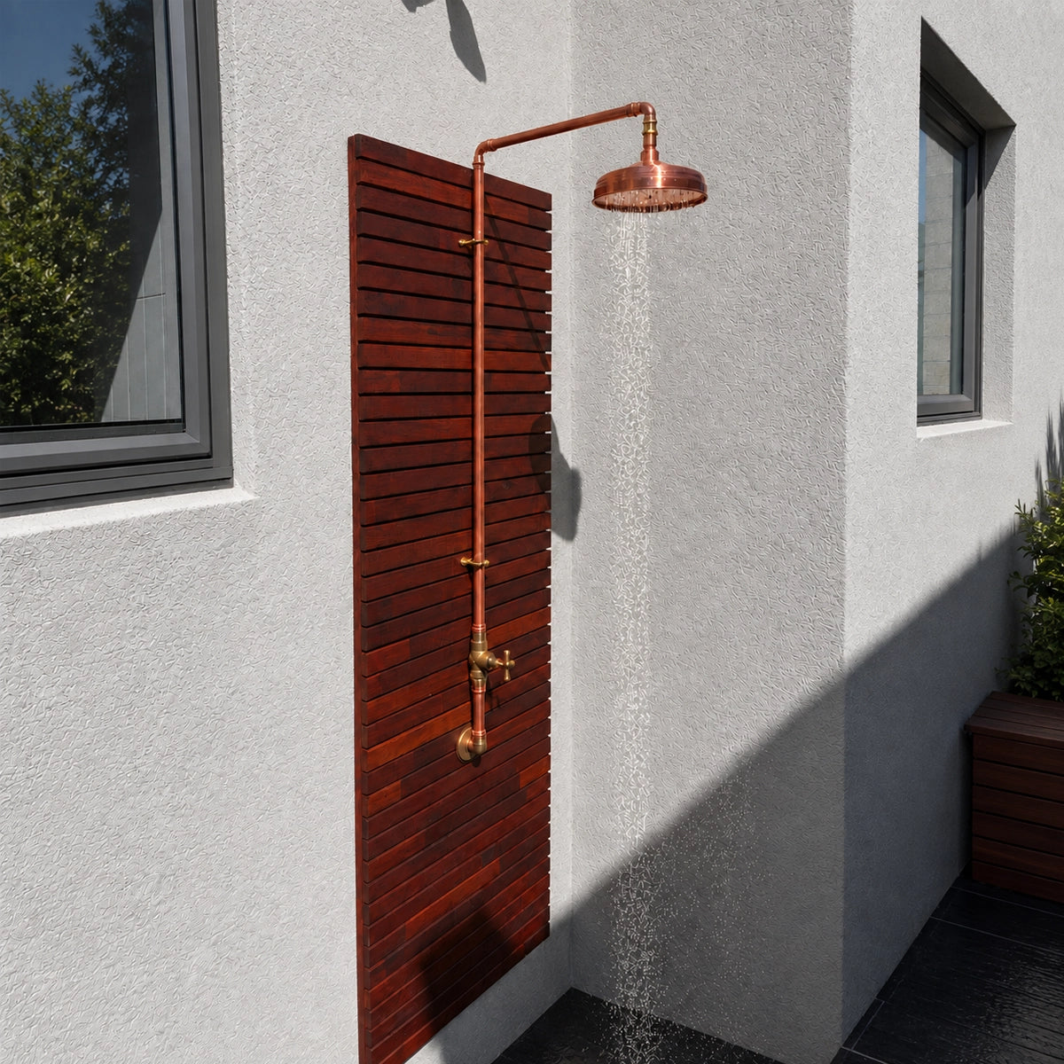 A copper outdoor shower system with a round shower head, vertical hose, and brass valve, mounted on a dark reddish-brown wooden slat panel against a light gray stucco wall.