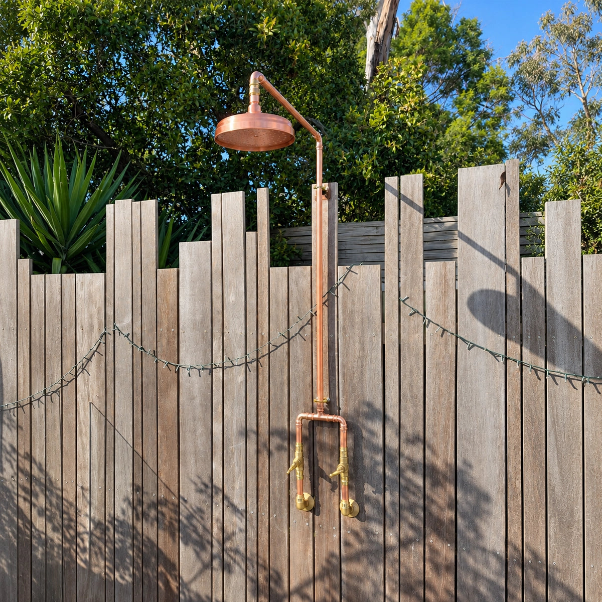 A copper outdoor shower system with a round shower head, curved arm, vertical pipe, and brass nozzles mounted against a wooden fence in a backyard setting with trees and sky behind it.