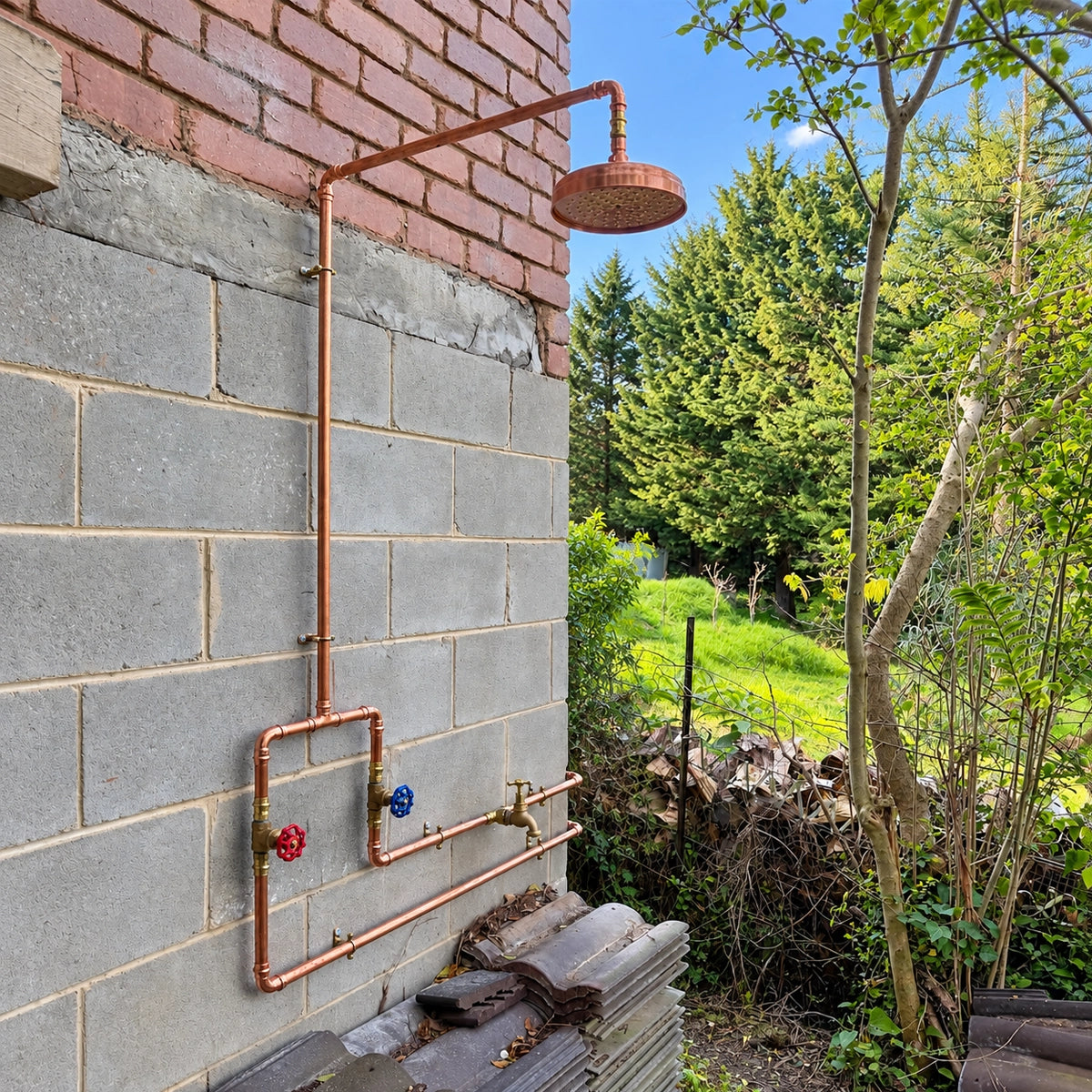 An outdoor wall-mounted copper shower system with a round shower head, copper pipes, and taps attached to a gray block wall, with trees and grass in the background.