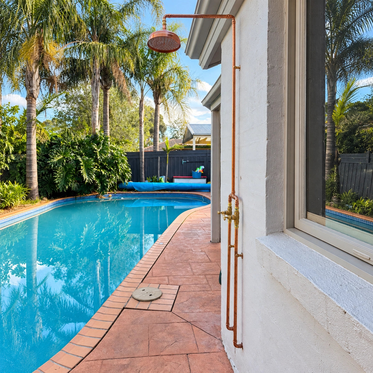 A copper outdoor shower system with a hanging shower head and brass faucet mounted on a white stucco house wall beside a swimming pool with palm trees in the background.